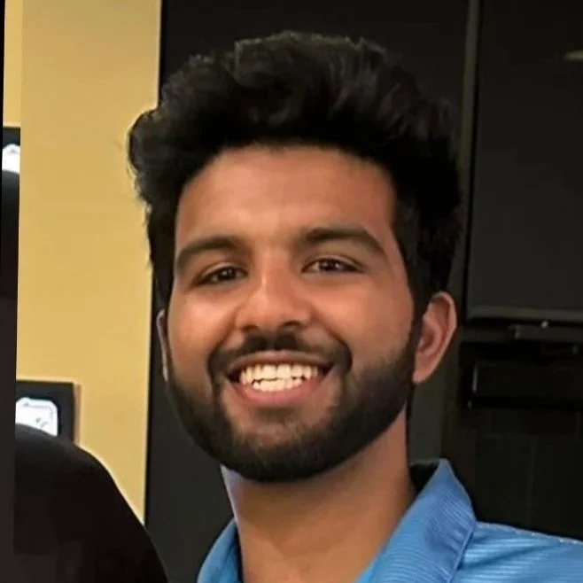 Smiling young man with dark hair and beard, wearing a blue shirt, indoors with dark background.