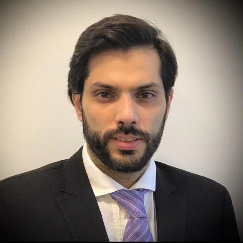 A man with dark hair and a beard wearing a black suit, white shirt, and purple tie, looking directly at the camera with a neutral expression.