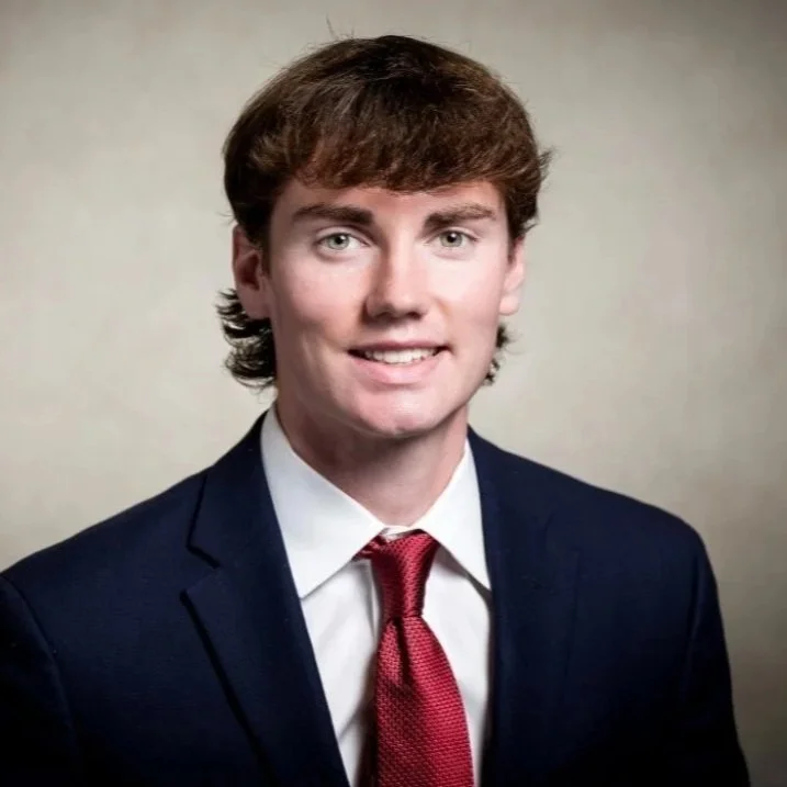A young man with light skin, brown hair, and wearing a dark blue suit, white shirt, and red tie, smiling for a formal portrait against a plain background.