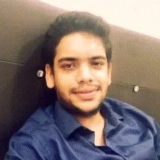 A young man with dark hair and a light beard smiling, wearing a blue shirt, sitting against a neutral background.