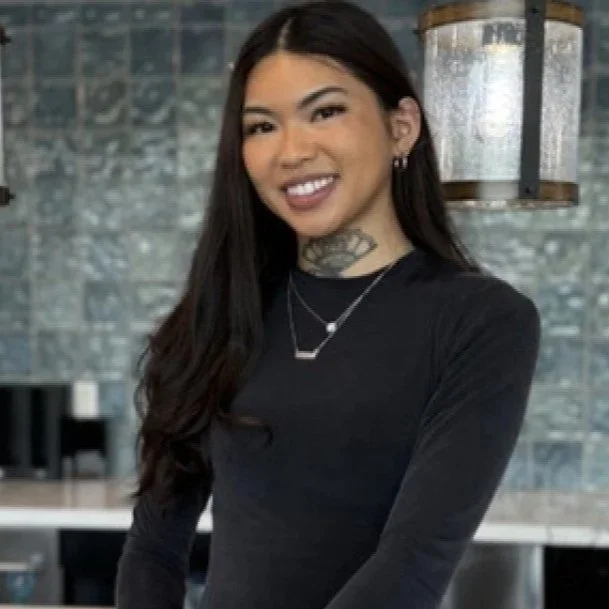 A woman with long dark hair, smiling and wearing a black long-sleeve top, jewelry, and earrings, standing in a modern kitchen with a textured grey backsplash.