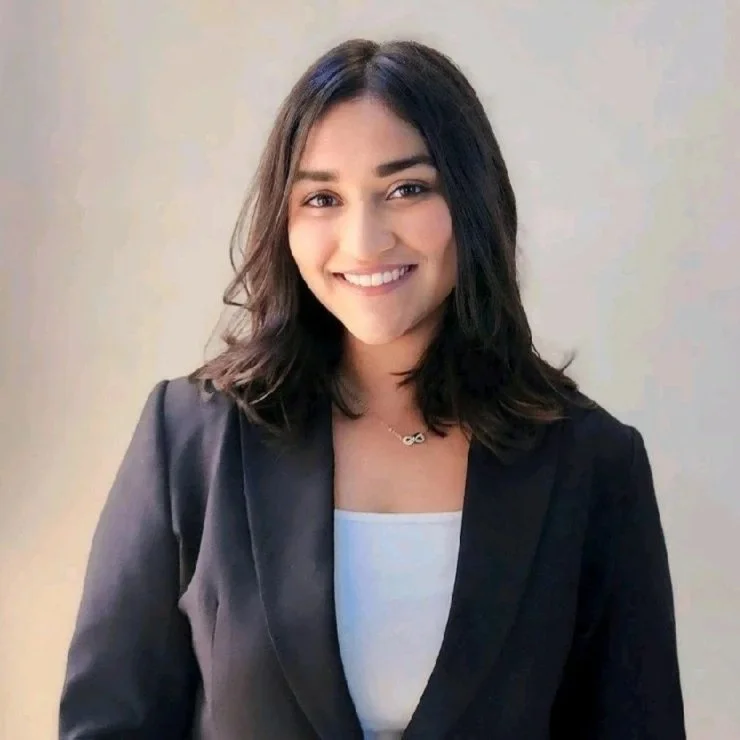 Young woman with shoulder-length dark hair smiling, wearing a black blazer and a white top, standing against a neutral background.