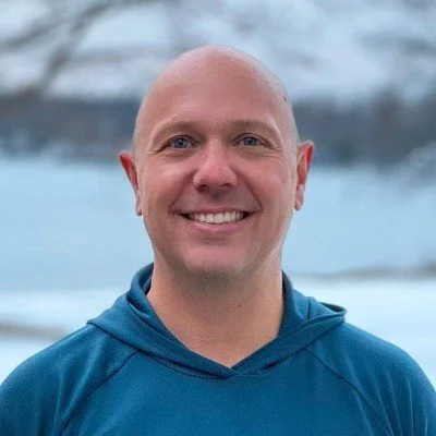 Portrait of a smiling man with a bald head outdoors near water and trees.