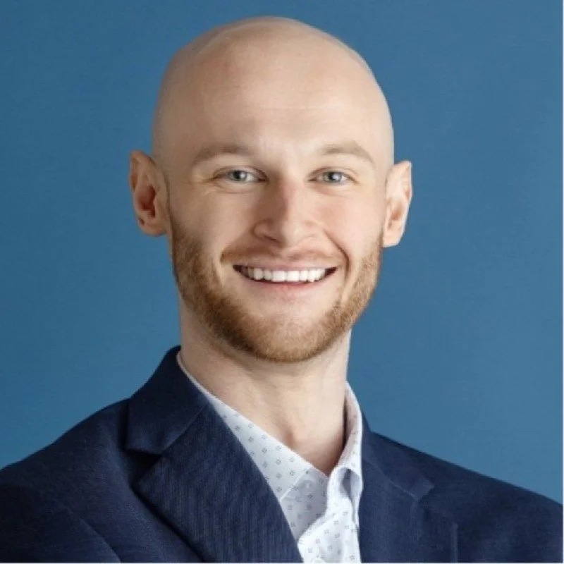 Headshot of a smiling man with a bald head and a beard, wearing a dark suit jacket and a white shirt with a subtle pattern, against a blue background.