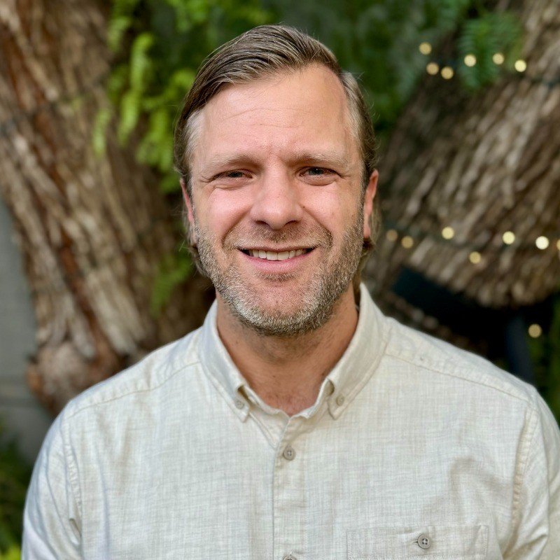 A middle-aged man with light skin, short light brown hair, and a beard, smiling outdoors in front of a large tree with string lights and green foliage, wearing a light-colored button-up shirt.