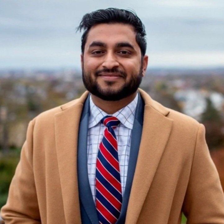 A man smiling outdoors in a tan coat, dress shirt, and red striped tie, with a blurred cityscape background.