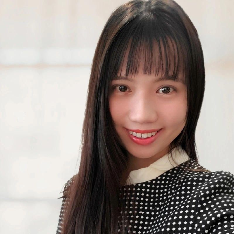 A young woman with long dark hair, bangs, and a bright smile, wearing a black polka dot top. She is inside a light-colored room.
