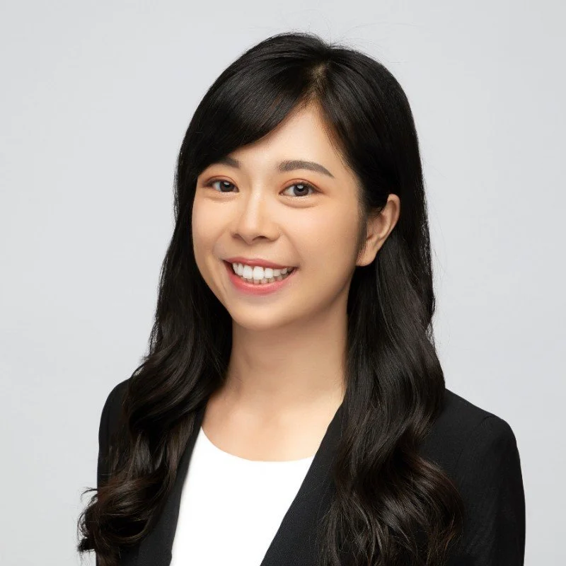 A woman with long black hair in loose waves, wearing a black blazer over a white top, smiling against a plain light gray background.