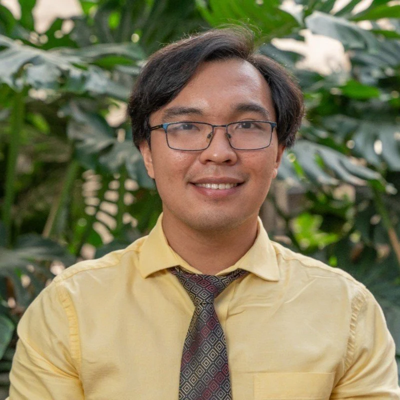 A young man with glasses and a yellow shirt, smiling outdoors with green foliage in the background.