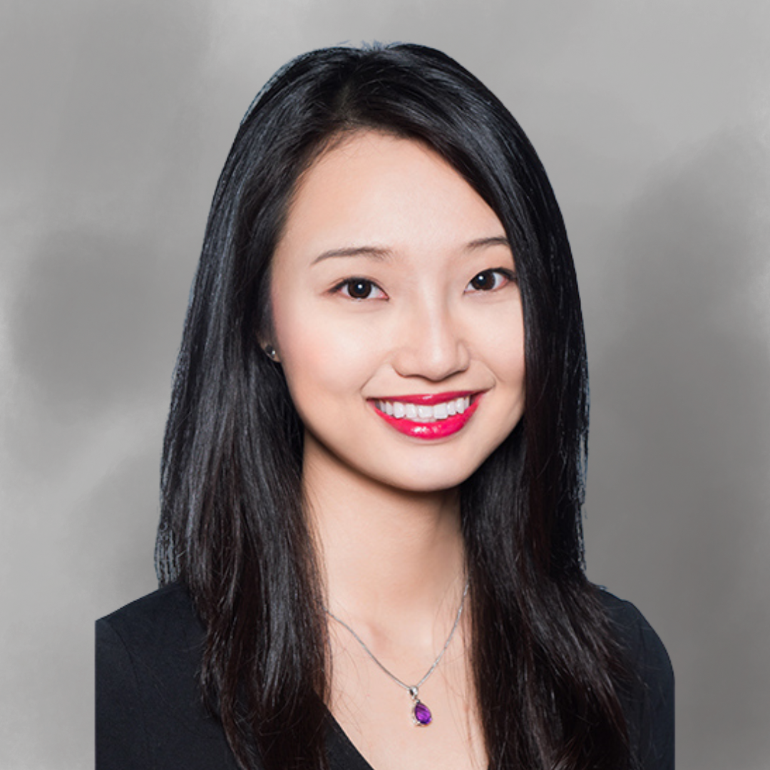 A woman with straight black hair wearing a black top, a necklace with a purple pendant, and red lipstick, smiling against a gray background.
