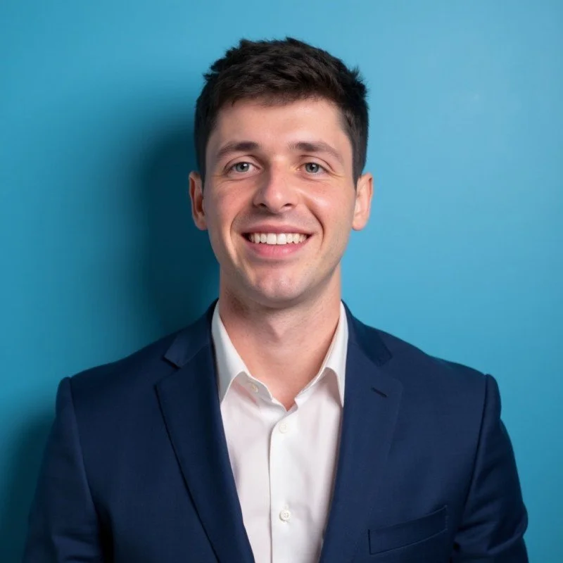A young man with dark hair, wearing a dark blue suit and white shirt, smiling, against a light blue background.