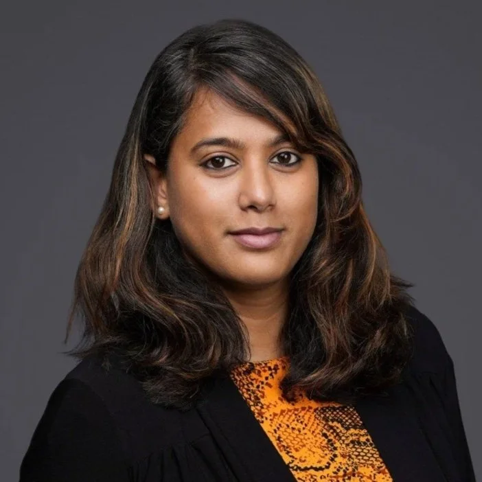 A woman with long, wavy dark hair and medium skin tone wearing a black judge's robe over an orange and black patterned top, against a gray background.