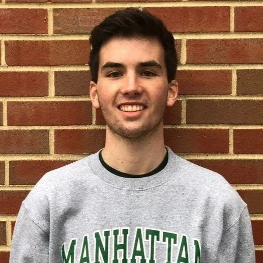 A young man with dark hair wearing a gray sweatshirt that says 'MANHATTAN' in green letters, standing in front of a brick wall, smiling at the camera.