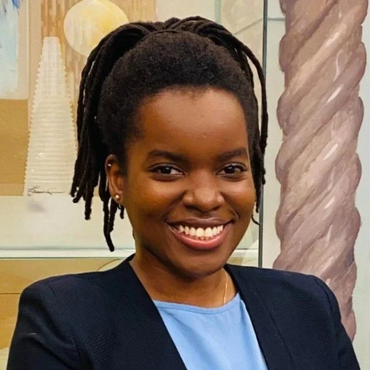 A young woman with dark skin and natural dreadlocks styled up, smiling and wearing a black blazer over a light blue top, standing indoors next to a large decorative twisted column.