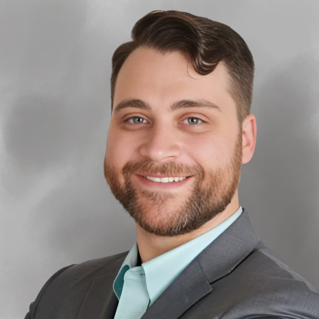 Headshot of a man with short brown hair, a beard, and blue eyes, wearing a dark suit and light blue shirt, smiling against a gray background.