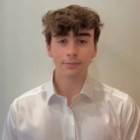 A young man with brown, curly hair wearing a white dress shirt, standing against a plain light-colored wall.