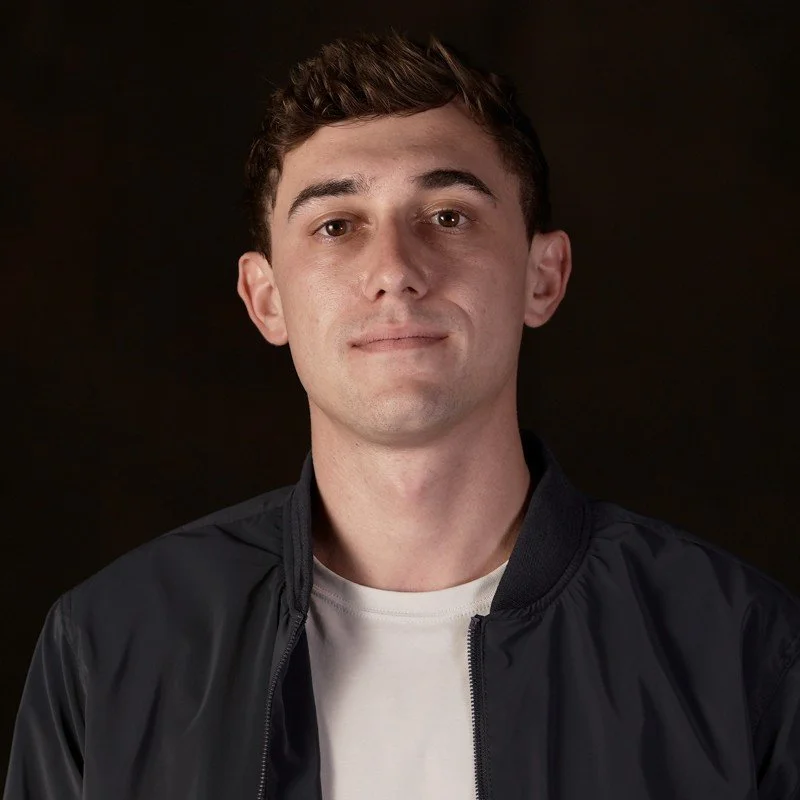 Portrait of a young man with short brown hair, wearing a black jacket over a white shirt, against a dark background.