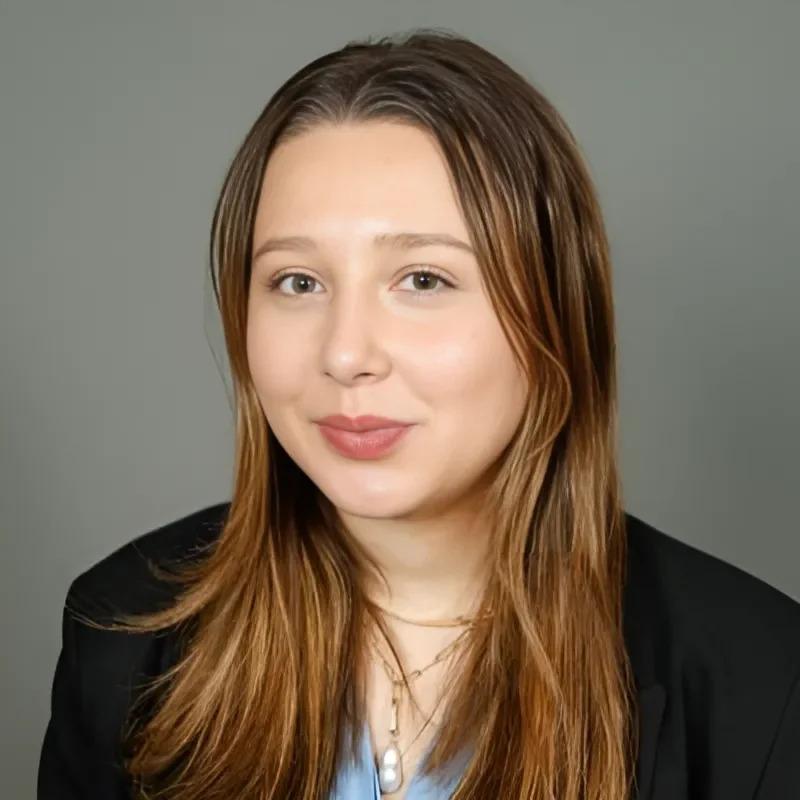 Portrait of a young woman with long, light brown hair, smiling softly, wearing a black blazer and layered necklaces, against a plain gray background.