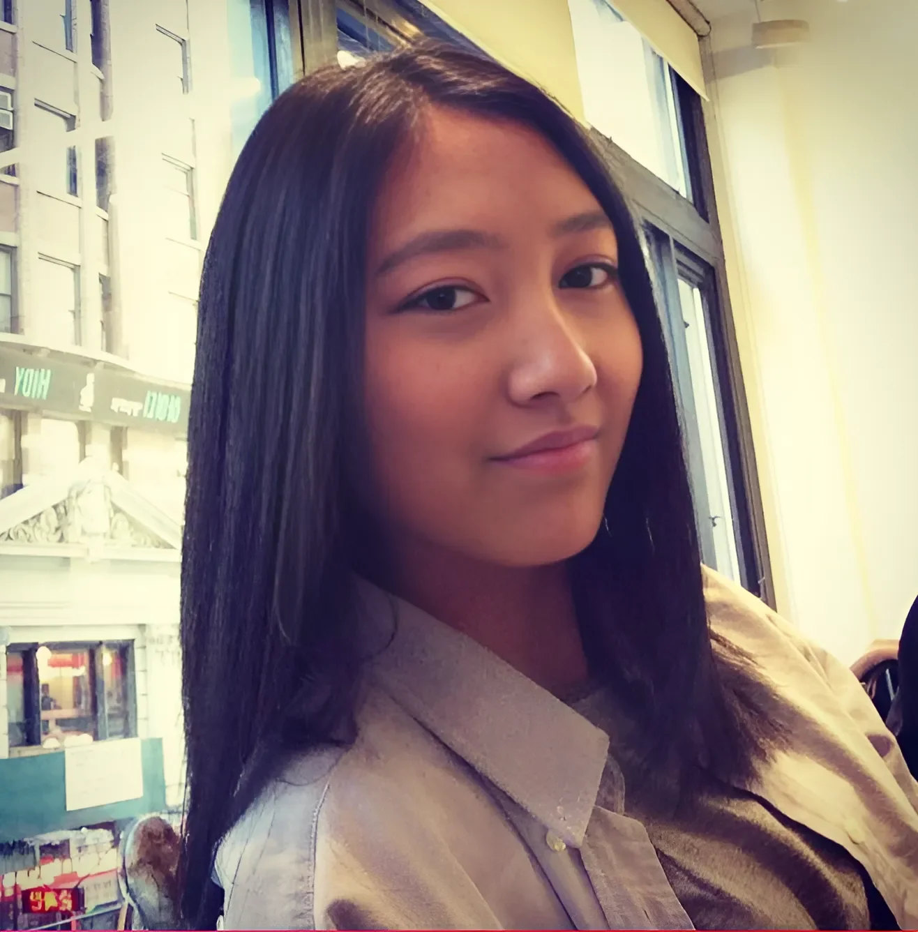 A young woman with straight black hair sitting by a window in a busy urban area, looking at the camera with a slight smile.