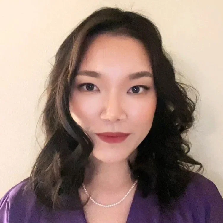 A woman with medium-length wavy dark hair and light skin, wearing a purple top and a pearl necklace, posing against a plain light-colored wall.