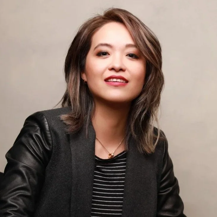 A young woman with shoulder-length brown hair styled in loose waves, wearing a black jacket with leather sleeves over a black and white striped shirt, and a small gold necklace, smiling slightly at the camera against a plain gray background.