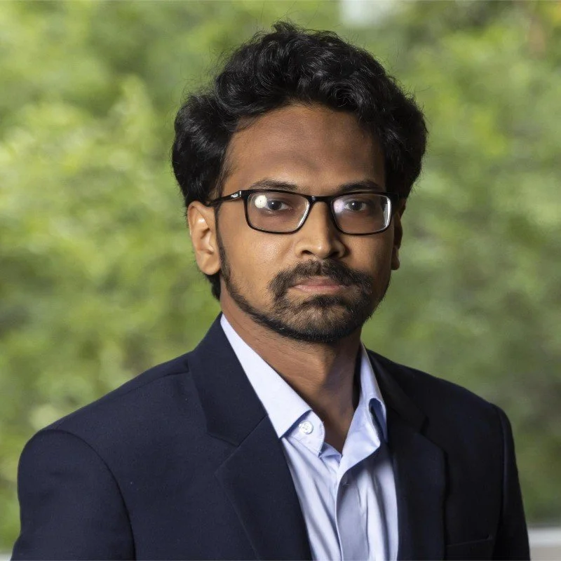 Portrait of a man with black hair, glasses, and a beard, wearing a dark suit and light shirt, set against a blurred green background.