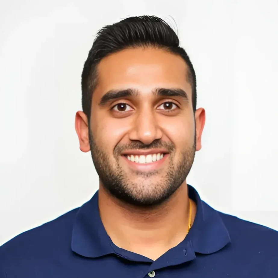 A smiling man with dark hair, facial hair, and brown eyes, wearing a navy blue polo shirt, standing against a plain white background.