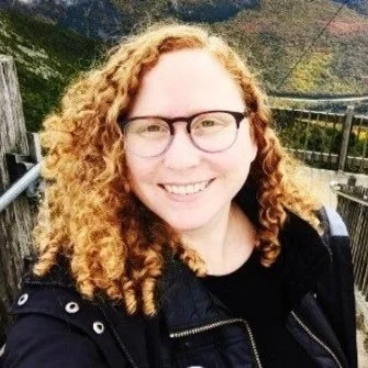 A woman with curly red hair and glasses smiling outdoors, with mountains and a railing in the background.