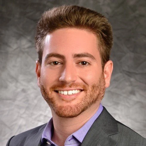 Portrait of a man with light brown hair and a beard, smiling, wearing a gray blazer and purple shirt, against a gray textured background.