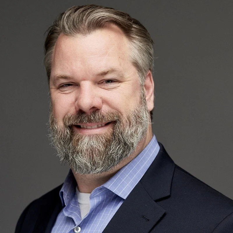 A middle-aged man with a beard and mustache, smiling, wearing a dark suit jacket over a light blue checkered shirt, against a gray background.