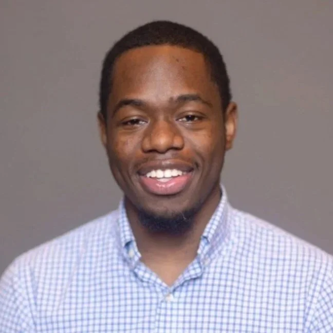 A young man with short black hair, light skin, and a slight beard smiling in front of a plain gray background, wearing a light blue checked button-down shirt.