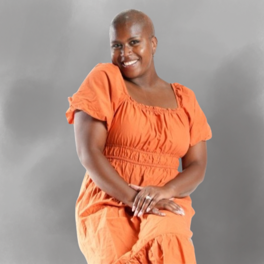 A woman with a shaved head, wearing an orange dress, smiling and sitting against a plain gray background.