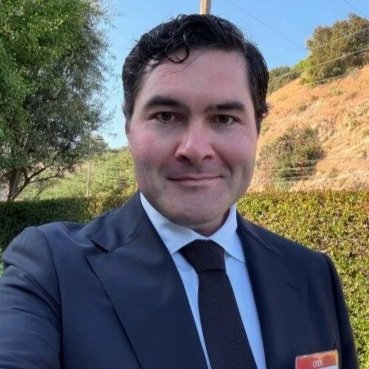 A man in a suit and tie taking a selfie outdoors with trees and hills in the background.