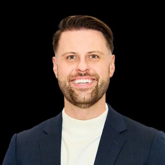 Smiling man with short hair and beard wearing a white turtleneck and navy blazer against a black background.