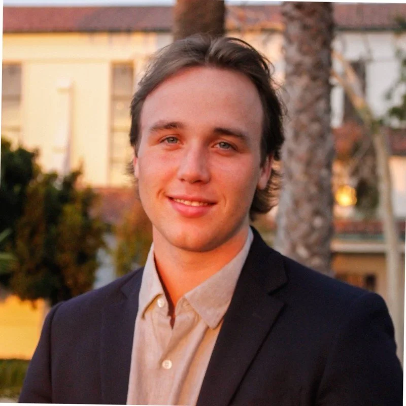 Young man with light brown hair smiling outdoors during sunset, wearing a beige shirt and dark blazer, with trees and a building in the background.