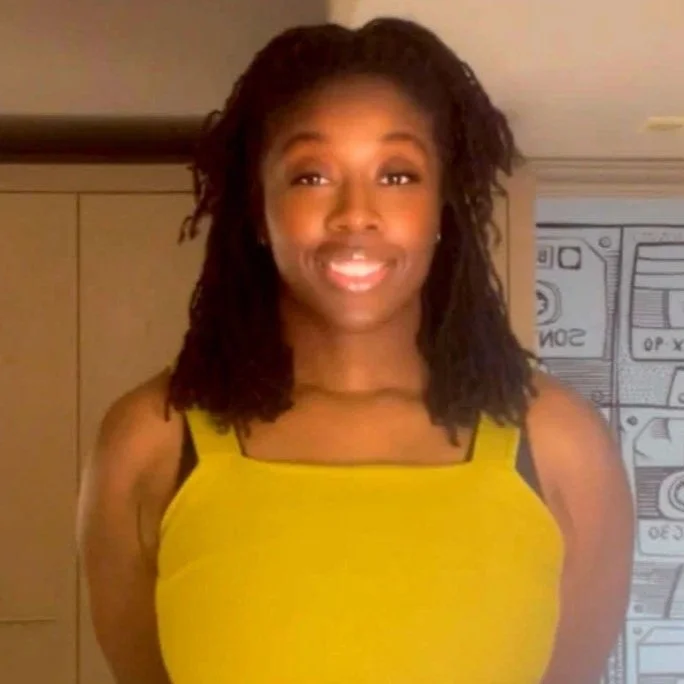 A woman with shoulder-length curly hair, wearing a sleeveless yellow top with black straps, standing indoors with a decorative wall in the background.