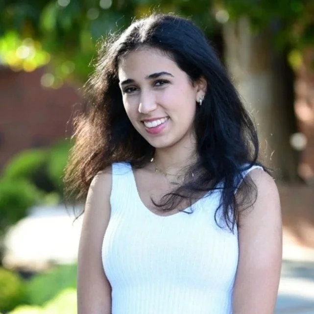 A young woman with dark hair and a white sleeveless top smiling outdoors with trees and greenery in the background.