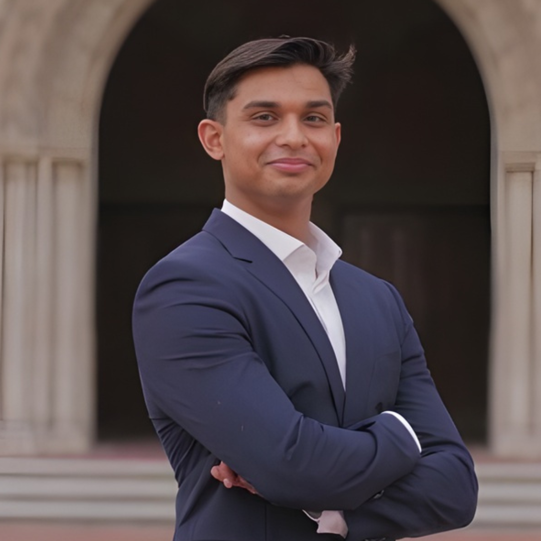 A young man in a navy blue suit standing outdoors with arms crossed in front of a stone archway, smiling at the camera.