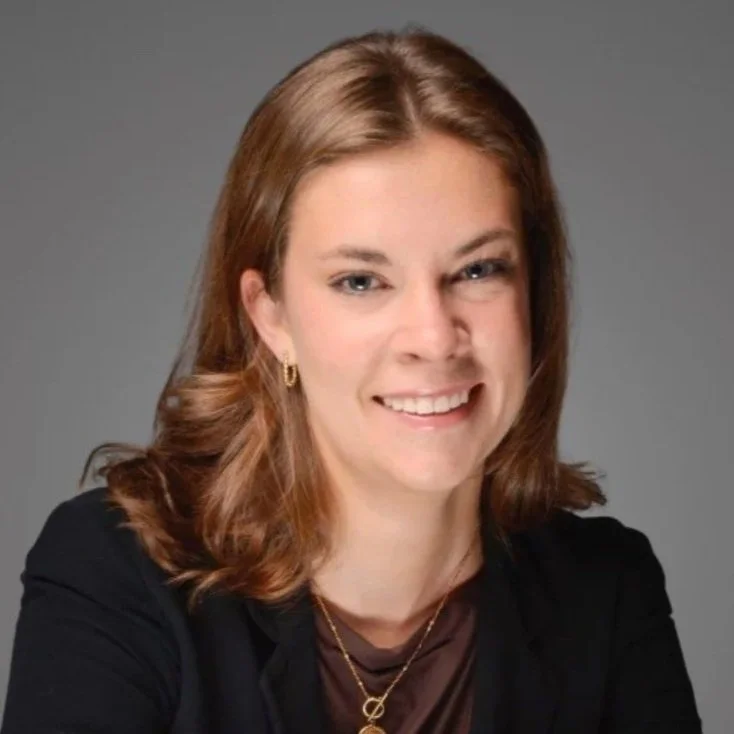 A woman with shoulder-length brown hair, wearing a black blazer and a gold necklace, smiling at the camera against a gray background.