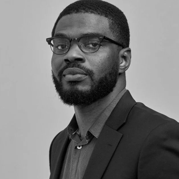 Black man wearing glasses, a blazer, and a patterned shirt, looking at the camera with a neutral expression.