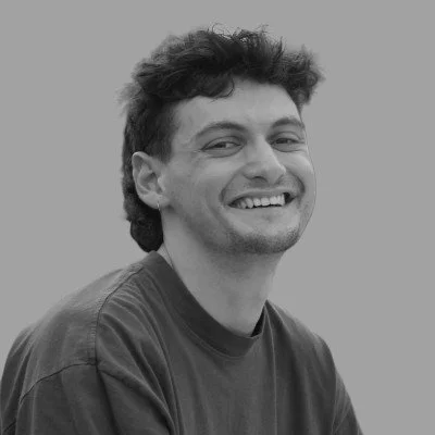 Young man with dark, curly hair smiling at the camera, wearing a casual T-shirt against a plain background.