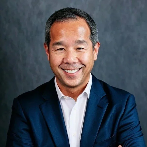 Smiling Asian man in a dark blue suit and white shirt against a gray background.