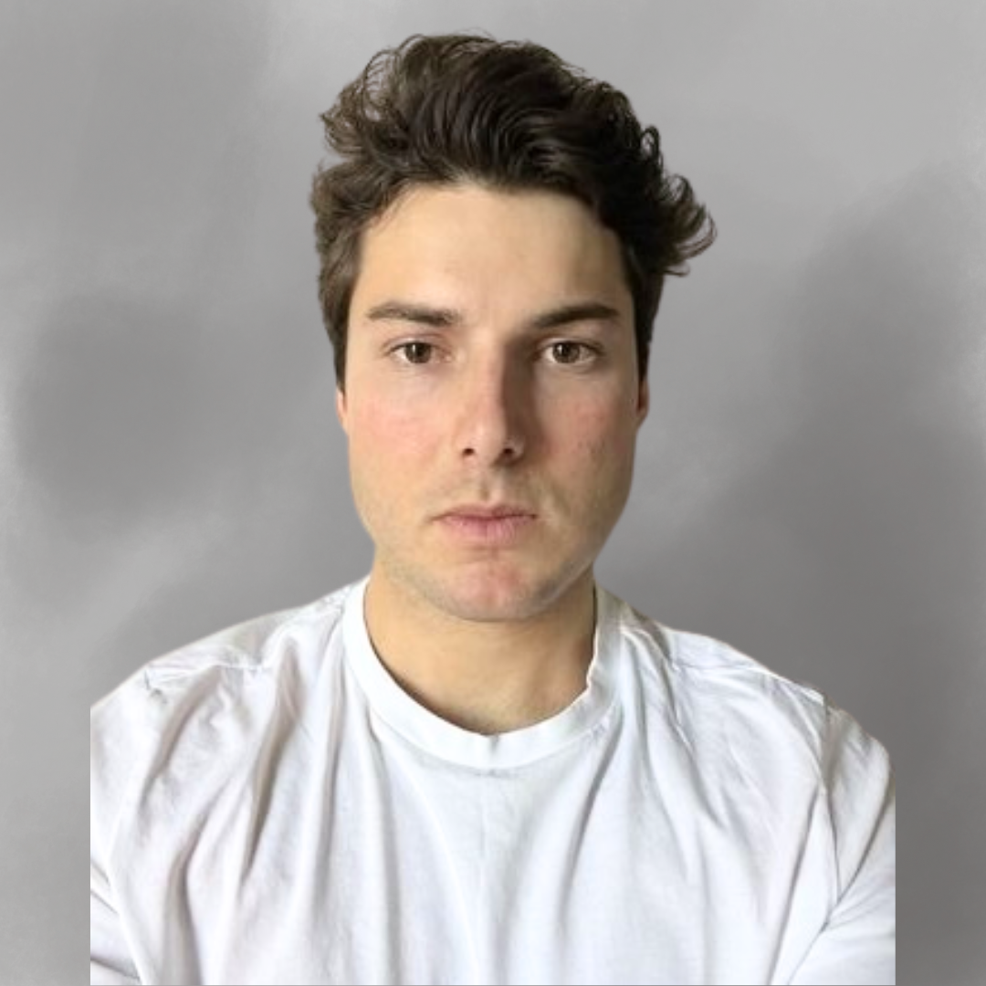 A young man with dark, wavy hair and fair skin taking a selfie against a plain gray background, wearing a white t-shirt.