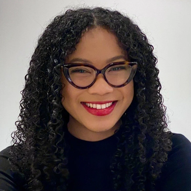 A young woman with curly black hair, glasses, and red lipstick smiling at the camera.
