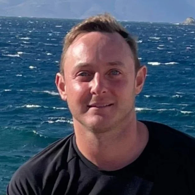 A man with short brown hair and fair skin standing in front of the ocean with waves and a cloudy sky.