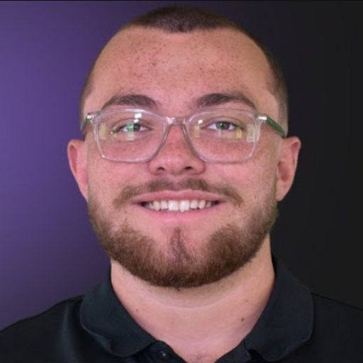 A young man with short hair, glasses, and a beard smiling at the camera against a dark background.