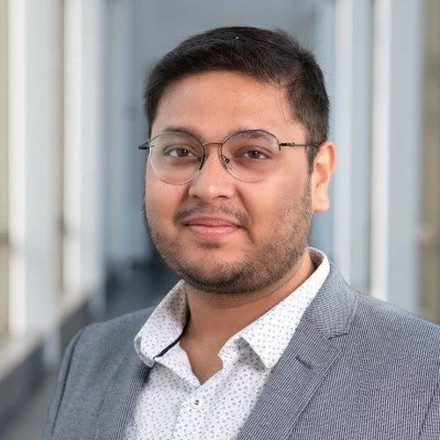 Professional headshot of a man with glasses, wearing a gray suit and white shirt, standing in an office environment.