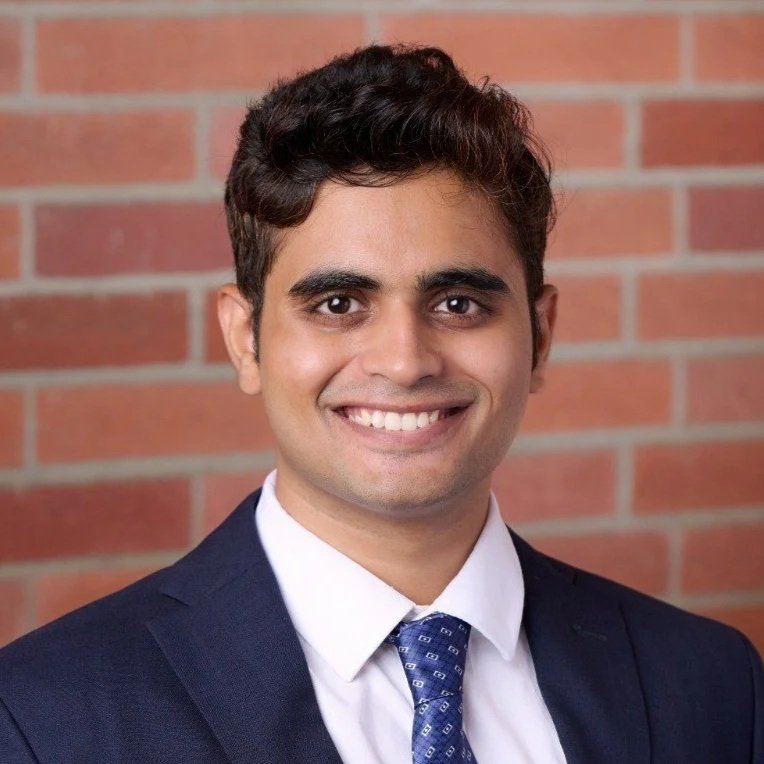 Headshot of a smiling man in a dark suit, white shirt, and patterned tie, standing in front of a brick wall.