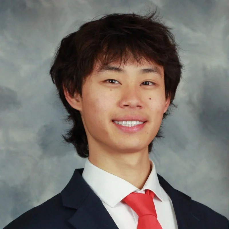 Young man with a smile, wearing a suit jacket, white shirt, and red tie against a cloudy background.