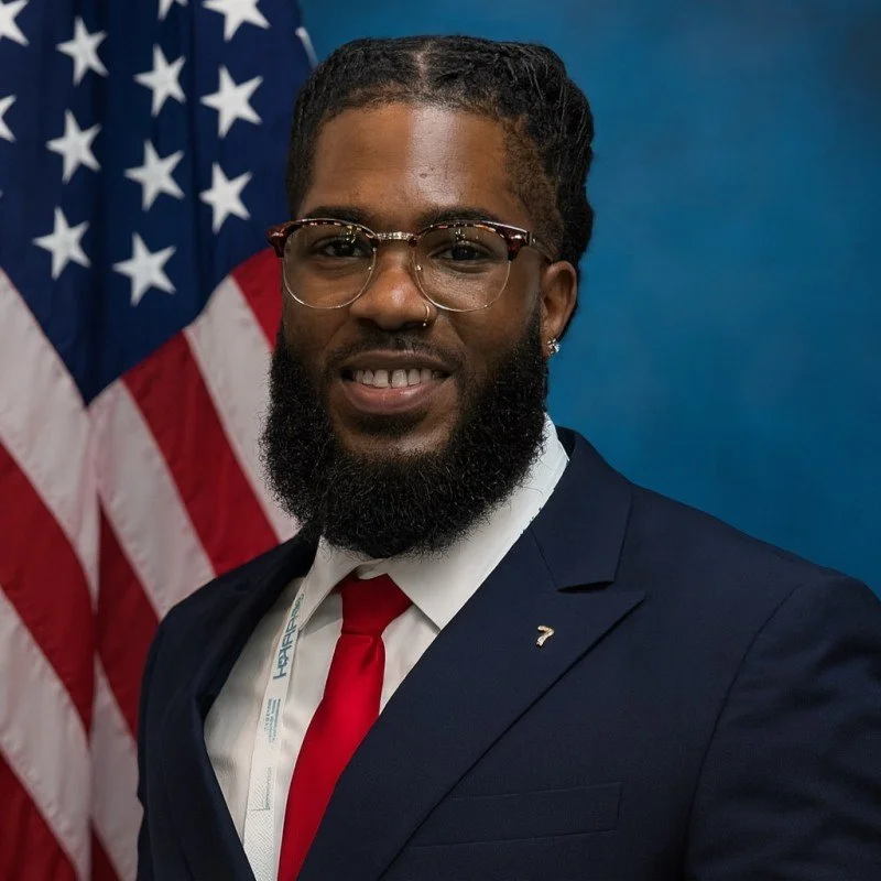 A man in a suit and red tie standing in front of an American flag.